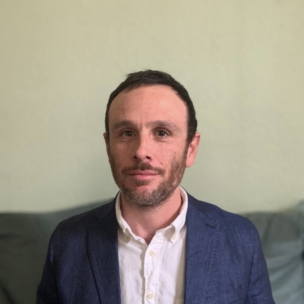 Photo from torso up of man with short hair and short beard wearing suit