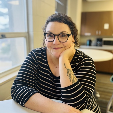 woman in striped long sleeved shirt, wearing glasses, sitting at table smiling, one hand on her cheek