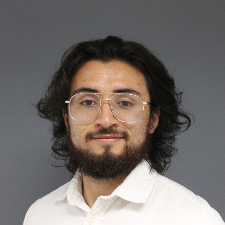 young man with longish brown hair and beard wearing glasses and white shirt in front of gray wall 